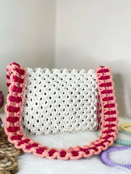 A lovely combination of colors, this white shoulder bag is accented with a strap woven from peach and pink cords. It’s a perfect accessory for spring and summer.