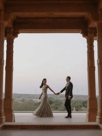 A classic, elegant pose within the pillared corridors of Suryagarh, Jaisalmer. The couple holds hands, their formal attire contrasting beautifully with the ancient stone and the expansive desert landscape visible beyond.