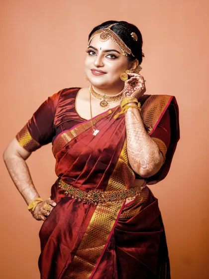 A classic bridal portrait against a simple orange background, highlighting the rich color of her maroon silk saree.