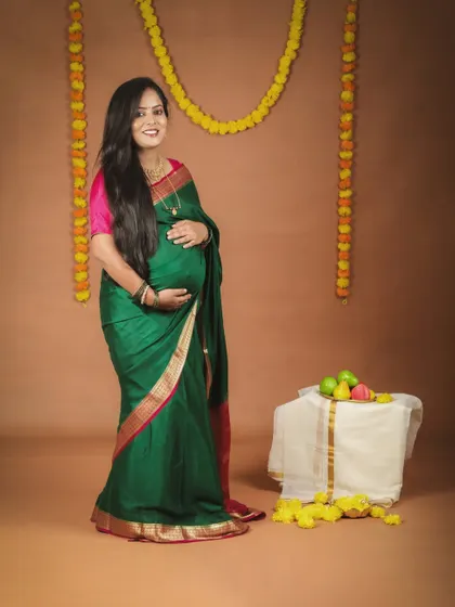 A full-length portrait of a smiling mom-to-be in a lovely green silk saree. The simple setup with marigold flowers creates a classic and traditional feel for her maternity photo.