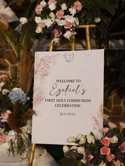 The welcome sign for Ezekiel's First Holy Communion. We designed it with a soft, rustic aesthetic using pampas grass illustrations and elegant script, surrounded by fresh flowers to greet the guests.