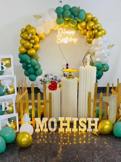 A cheerful first birthday setup for Mohish, with a green and gold balloon ring, a picket fence, and 'BABY' blocks.