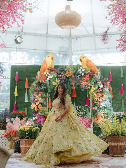 A stunning bride twirling in her vibrant Mehendi outfit. The backdrop of a floral-covered gazebo with colorful parrot motifs and tassels perfectly complements her joyful energy.