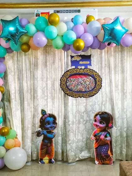 A naming ceremony setup at home with a pastel-colored balloon arch. The theme includes Radha Krishna cutouts and transparent 'BABY' blocks.