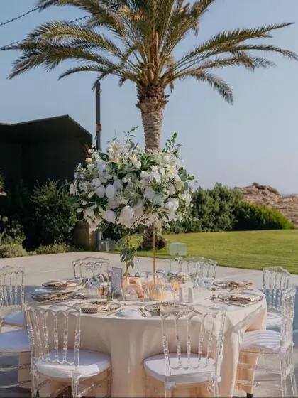A simple and elegant outdoor table setting. The clear ghost chairs and a single, lush floral centerpiece create a clean and modern look against the natural backdrop.