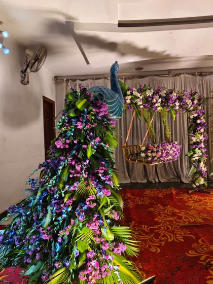A close-up of a peacock-themed decor piece for a naming ceremony. This peacock is crafted with a mix of purple and blue flowers, creating a vibrant and artistic focal point.