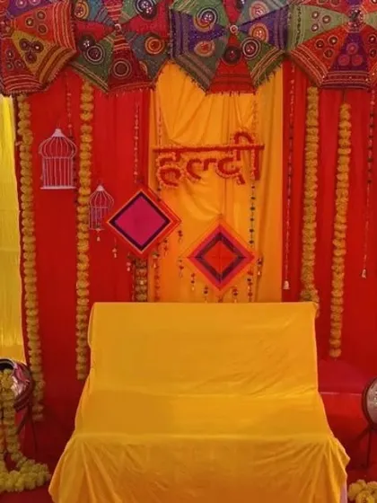 A traditional Haldi seating area with a backdrop of red and yellow drapes, decorated with umbrellas and marigold-filled pots.