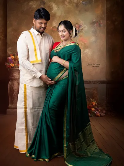 A gorgeous maternity portrait featuring a deep green saree. The couple's pose and the classic studio backdrop are inspired by the timeless beauty of Ravi Varma paintings.