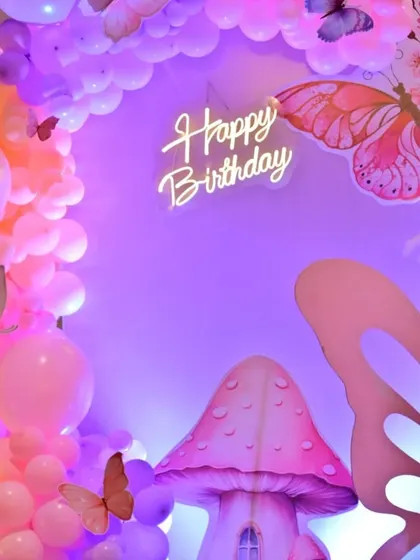 A close-up of the butterfly party backdrop. The soft purple lighting, neon sign, and detailed props like the mushroom house create a whimsical and enchanting atmosphere.