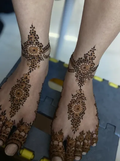 A matching pair of Arabic-style foot mehendi. The design is light and elegant, with a central trail from the toes to the ankle, adorned with delicate chains and mandalas.