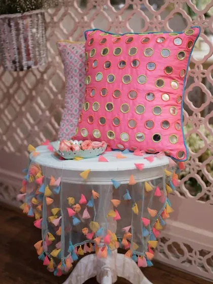 A pink cushion with mirror work and a small table with a colorful tassel tablecloth. These details are perfect for adding a touch of Indian craftsmanship to Mehendi or Diwali parties.
