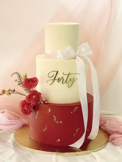 An elegant three-tier cake for a 40th birthday. The design features a bold red bottom tier with gold leaf, a simple white middle tier with a "Forty" script, and a smaller top tier, all tied together with a white ribbon.