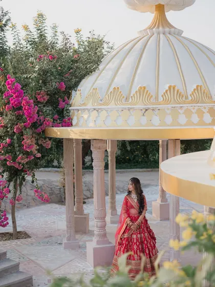 The bride in her red lehenga stands in a picturesque white and gold gazebo, surrounded by the lush pink bougainvillea of a Jaipur garden.