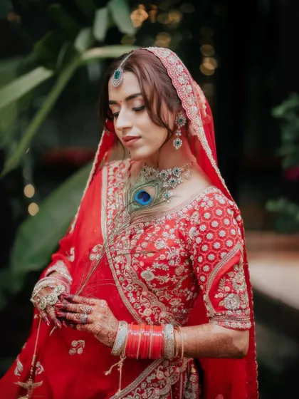 A portrait of elegance amidst nature. The bride, in her stunning red lehenga, is captured in a moment of quiet grace, the peacock feather in her jewelry adding a touch of royalty.