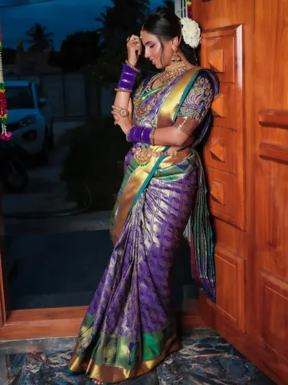 A full-length portrait of a model in a stunning purple silk saree, leaning against a doorway. The evening light and her elegant pose create a moody and sophisticated atmosphere.