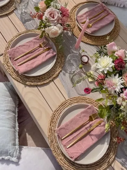 A closer look at a rustic-chic picnic table setting. The combination of natural wood, woven placemats, and soft pink linens creates a relaxed yet elegant feel.