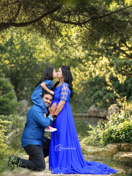 A heartwarming outdoor family portrait where the whole family is coordinated in shades of blue. The daughter, sitting on her father's shoulders, leans in to kiss her expectant mother, perfectly capturing the excitement of a growing family amidst a serene, natural setting.