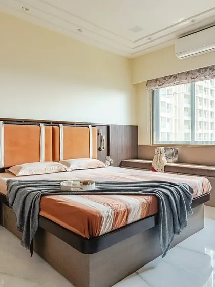 This bedroom is designed with warm, earthy tones, featuring an orange upholstered headboard against a dark wood-paneled wall. A custom window seat with built-in storage provides a cozy spot to relax and enjoy the view.