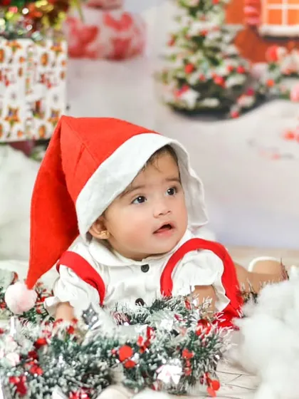 A cute baby dressed as Santa's little helper sits in a festive Christmas-themed setup, complete with presents and snow, ready for a holiday mini-session.