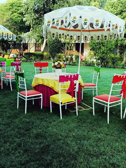 A fun and colorful outdoor seating area with multi-colored chairs and a block-printed umbrella, ideal for a Mehendi mela.