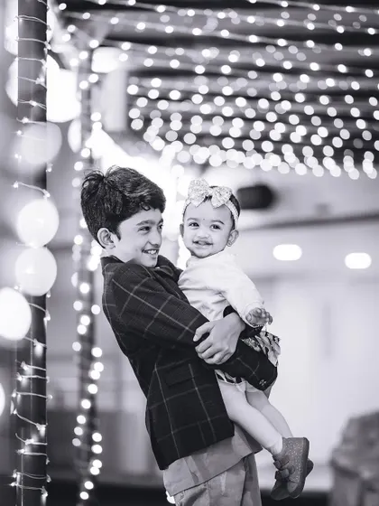 A brother holds his baby sister, both smiling, against a backdrop of beautiful bokeh lights. This black and white portrait emphasizes their sweet connection.