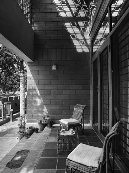 This black and white photograph of the Reshma Residence veranda emphasizes the building's architectural form and the play of light and shadow. The strong geometric patterns created by the sun filtering through the structure become a key design element.