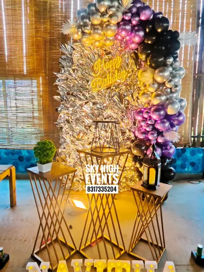 A chic surprise party setup with a silver tinsel backdrop and a black, purple, and silver balloon arch. The gold pedestals add a touch of modern elegance.