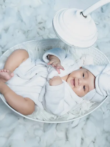 This happy baby is lying in a white bowl surrounded by soft cotton clouds, looking like a little angel.