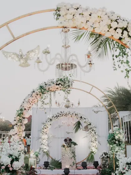 A beautiful all-white wedding setup with grand circular arches and crystal chandeliers. The lush floral arrangements add a touch of romance and elegance to the outdoor ceremony.