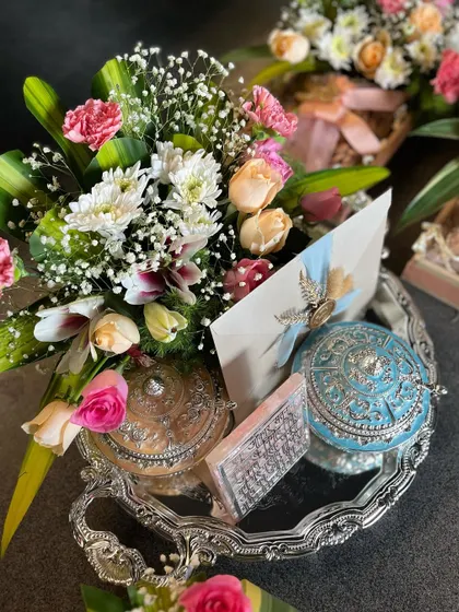 This Samdhi invitation set includes a silver tray with ornate, hand-painted boxes and a custom-sealed invitation card, all surrounded by a beautiful arrangement of fresh flowers.