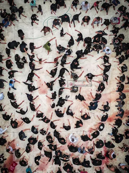 A closer top-down image of a Muharram ritual, focusing on the abstract patterns created by the participants. The stark contrast of black clothing against the ground creates a powerful and emotional visual story of faith and remembrance.