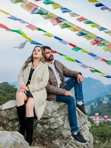 A couple sits on a rock with colorful Tibetan prayer flags fluttering above them, set against a backdrop of the Mussoorie hills.
