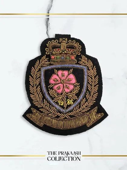 A beautiful hand-embroidered crest patch featuring a pink flower at its center, surrounded by a gold wreath and banner. This detailed appliqué is ideal for personalizing blazers or school-inspired fashion.