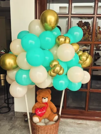 A unique "hot air balloon" decoration for a welcome baby party. The basket holds a cute teddy bear, and the balloon is made from a mix of teal, white, and gold balloons.