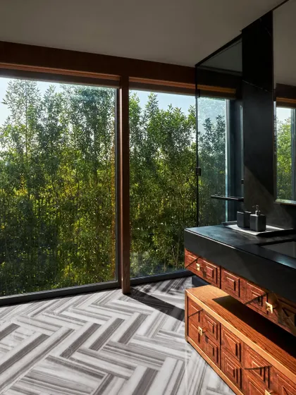 This bathroom at the Baid Residence is a study in natural textures, with a large window framing a view of the forest. The flooring is Equador stone, and the custom vanity combines wood with a black granite counter, creating a serene, spa-like feel.