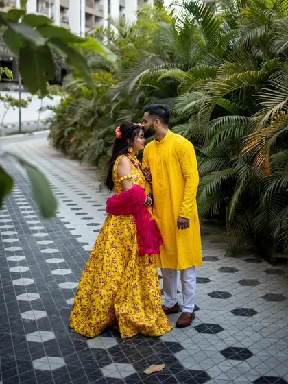 A full-length portrait of a couple in coordinated yellow outfits for their Haldi ceremony, sharing a tender moment in a lush garden setting.