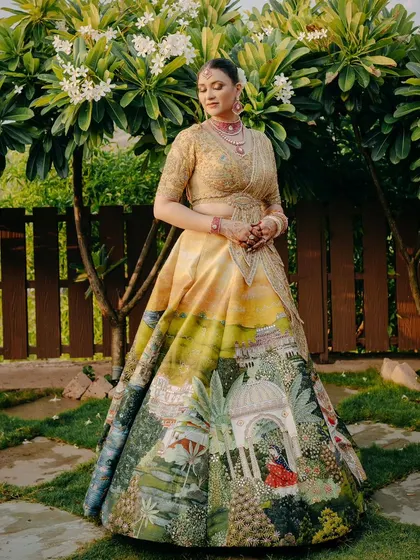 The bride stands in a garden, her unique lehenga featuring a painted landscape that tells a story. This showcases how I work with couples to integrate personal style into every aspect, including their wedding attire.