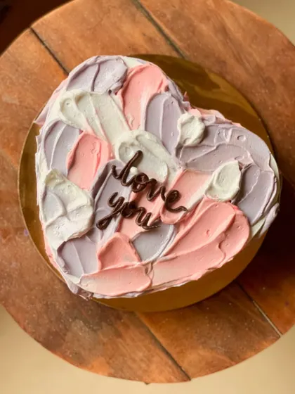 A lovely heart-shaped cake with a textured, painterly frosting effect in shades of pink, purple, and white. A simple "love you" message makes it perfect for any romantic occasion.