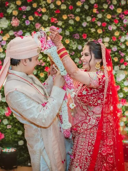 The playful Varmala exchange. The bride laughingly places the garland on her groom, set against a vibrant wall of flowers.
