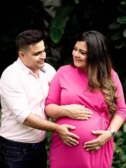 A classic couple's pose against a lush, dark green background. The contrast makes the mom-to-be's bright pink dress stand out, highlighting her and the baby bump beautifully.
