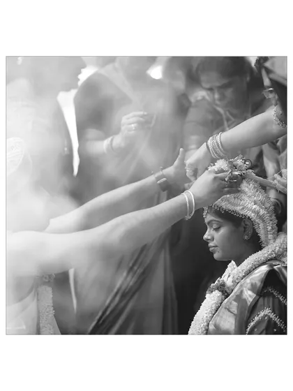 A black and white photo capturing the details of a Seemantha ritual. The image focuses on the hands placing a traditional floral headpiece on the mother-to-be, surrounded by smoke from the ceremony.