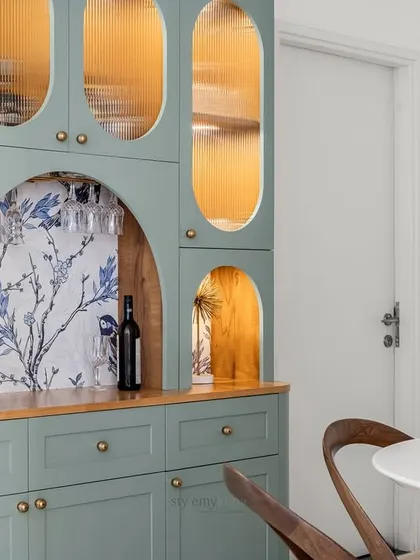 A close-up of a beautifully crafted crockery unit, featuring sage green cabinetry with curved details and fluted glass. The arched niche with bird-patterned wallpaper adds a whimsical and artistic touch to this functional piece.