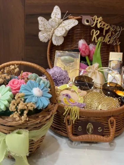 A duo of Rakhi hampers. One is a mini bouquet of soap flowers, and the other is a larger basket filled with sunglasses, a sparkly clutch, and other goodies for a stylish sister.