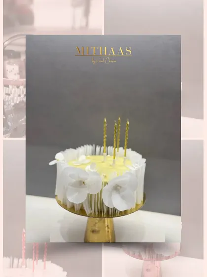 An elegant and minimalist celebration cake. This one is adorned with delicate white edible flowers and shimmering gold candles, presented against a sophisticated grey backdrop.