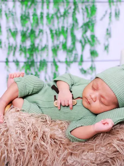 A newborn in a sage green outfit rests on a fluffy, textured prop. The backdrop of hanging greenery adds a fresh, botanical element to the studio setup.