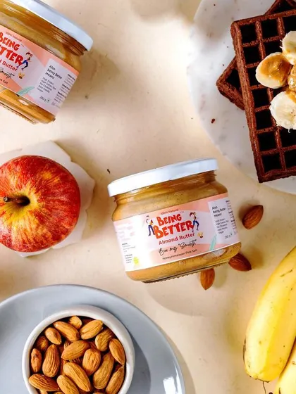 A bright and cheerful flat lay of Being Better's almond butter. The product is surrounded by its core ingredients like almonds, apples, and bananas, alongside waffles, visually communicating its healthy and delicious nature.