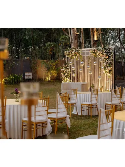 A wider view of the Sangeet setup, with elegant round tables and Tiffany chairs arranged on the lawn, ready for a night of celebration under the stars.