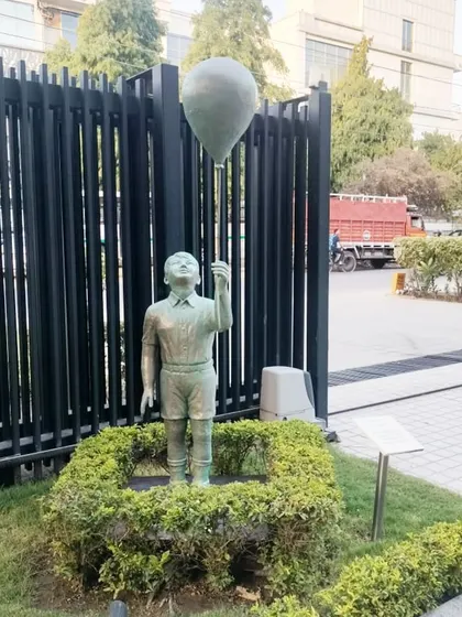 A charming life-size sculpture of a young boy holding a balloon, installed in a garden in Gurgaon. The piece has a classic weathered bronze finish.