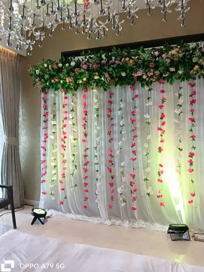 An elegant backdrop for a home party. A sheer white curtain with hanging pink and white flower strings is topped with a lush green and pink floral arrangement.
