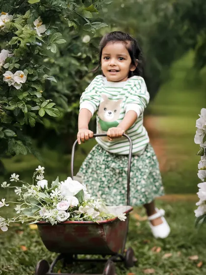 Playtime in the garden. Pushing a little wheelbarrow full of flowers, this little one is all smiles, creating a lively and adorable outdoor portrait.